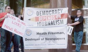 A still image from Stripped From Parts of journalists protesting. Two hold up a large sign with "Democracy Depends on Journalism" and "Heath Freeman, Destroyer of Newspapers."